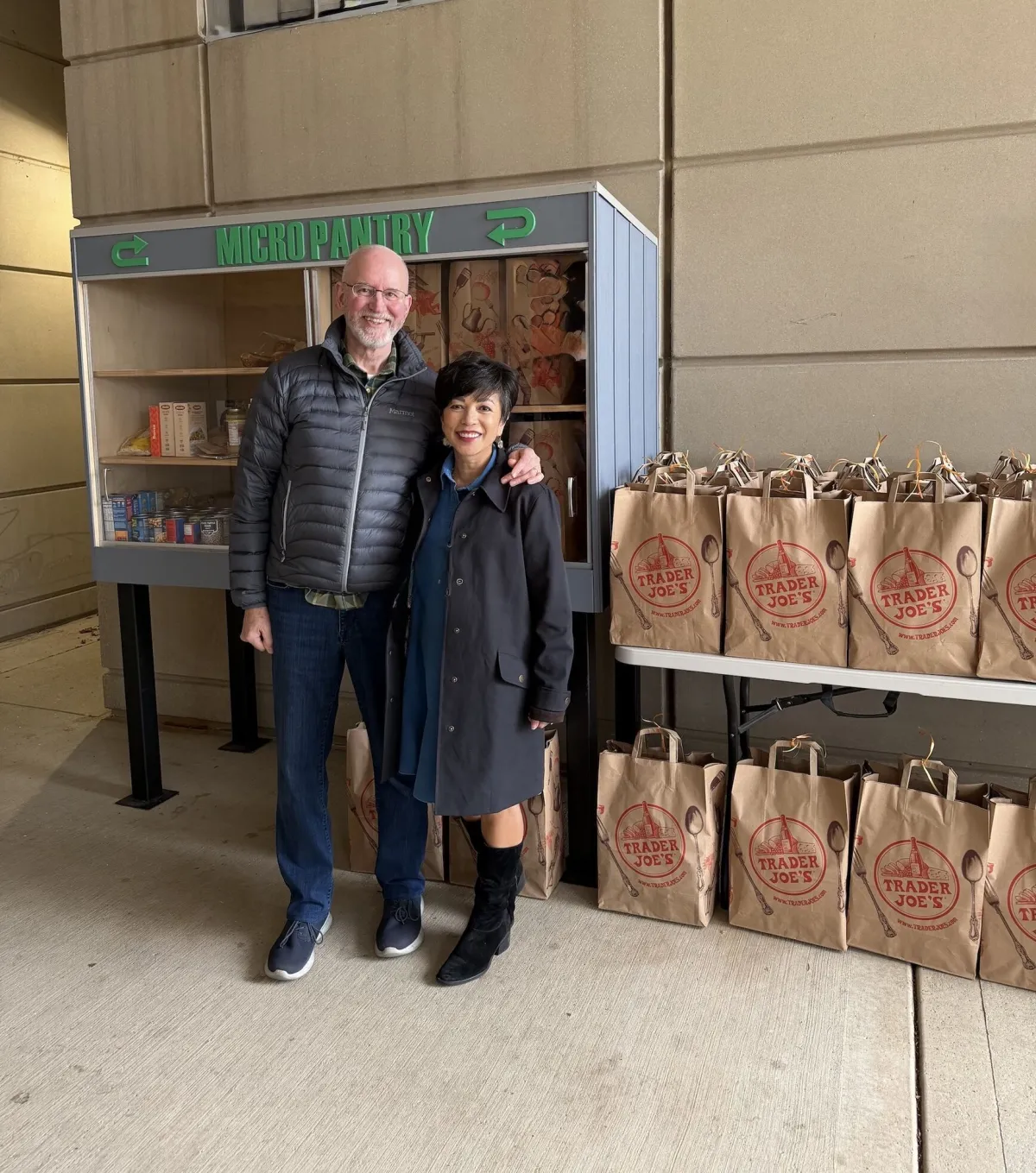 A man and a woman pose for a photo in front of a micro food pantry.
