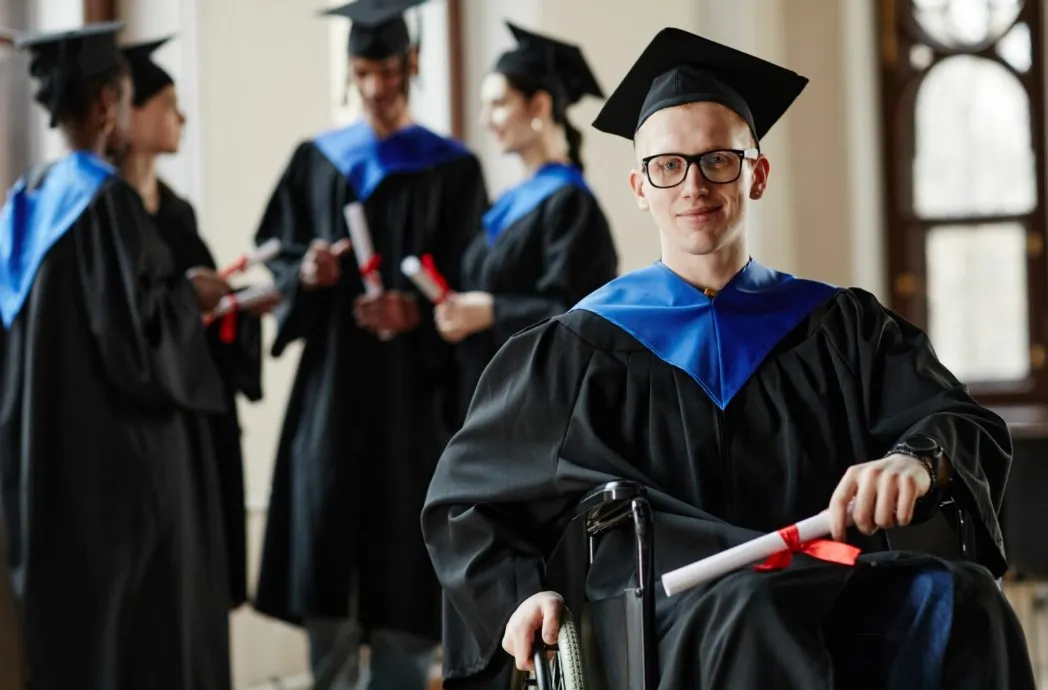 A student in a wheelchair wears a graduation cap and gown and holds a diploma.