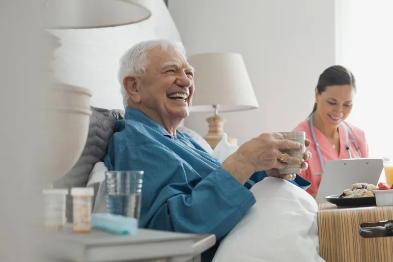 An elderly man seating on his bed in company of a nurse