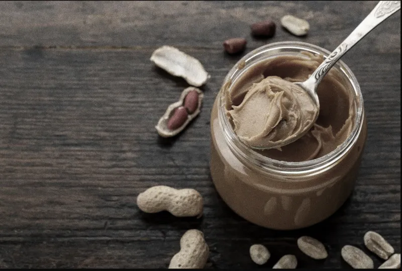 A spoonful of peanut butter sitting on a jar of peanut butter and peanuts surrounding the jar on a table.